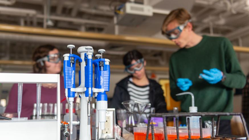 Students in protective gear discuss an experiment with scientific instruments in the foreground.