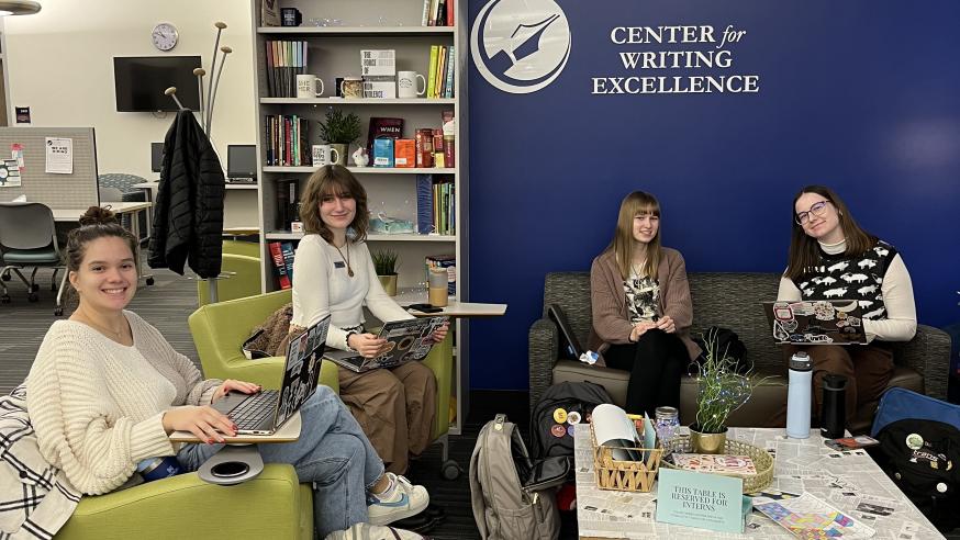 Four female presenting students are sitting on chairs and couches in front of a blue wall, which has the logo for the Center for Writing Excellence on it in white. Three of the students have their laptops open. They are all smiling.