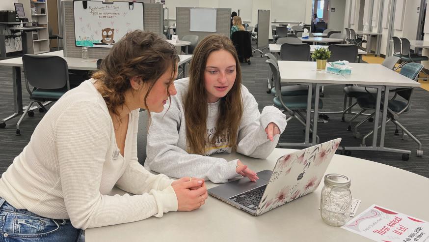 Two female presenting students are sitting at a round table. They are both leaning forward looking at a laptop.