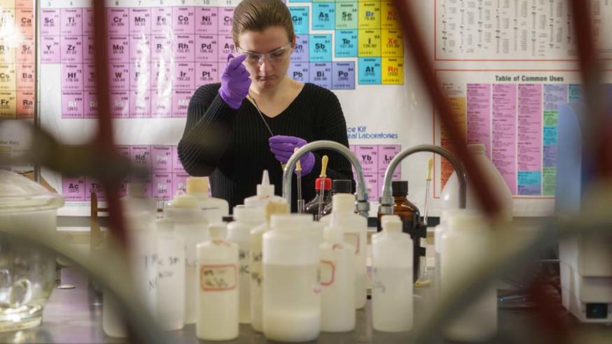 A student works with lab instruments with the periodic table hanging on the wall behind.