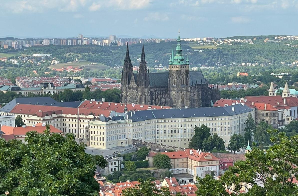 An aerial view of Prague showing the historic Prague Castle with its Gothic spires, surrounded by modern cityscape and lush greenery.