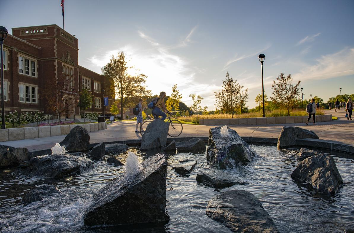 Students walk and bike next to the Stowe Fountain during a sunset on campus