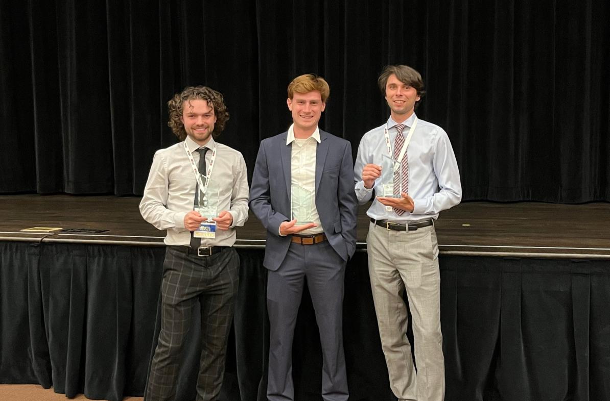 3 male students standing holding awards near a stage