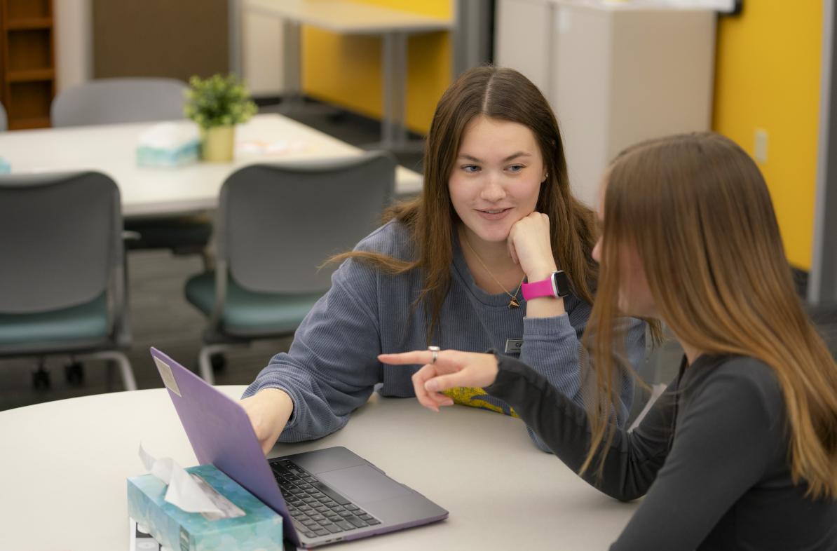 two female students at a table for a tutoring sessions, laptops on the table, both are looking at the screen