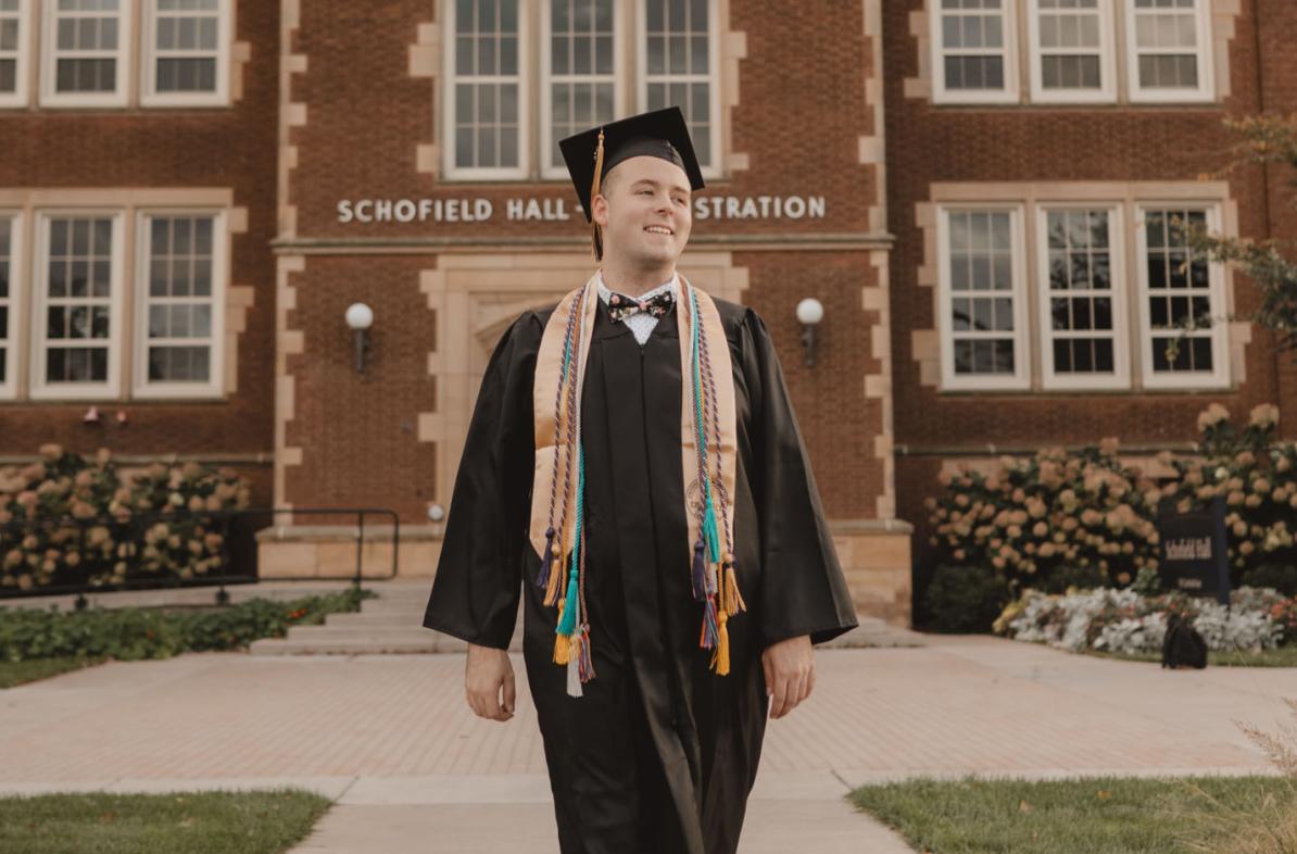 Jordan Blue smiling  in cap and gown in front of Schofield Hall, summer or fall sunset hour