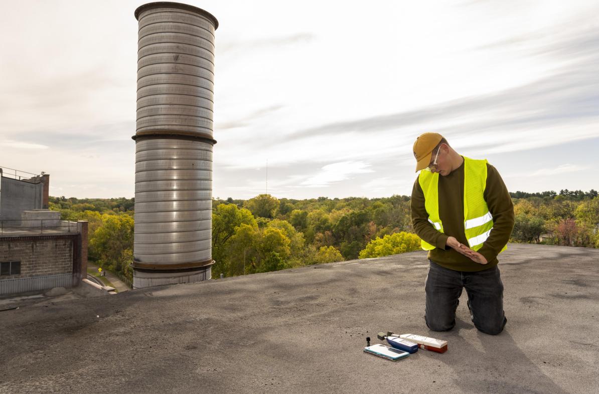 A male student wearing safety gear kneeling on a metal rooftop checking some measurement instruments