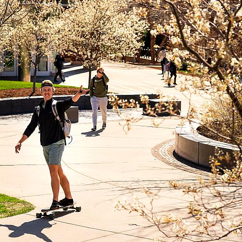 male student on a long board crossing campus