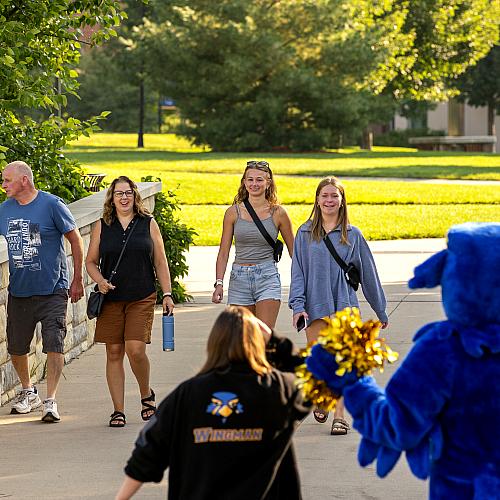 students and family crossing campus mall bridge facing Blu with spirit gear